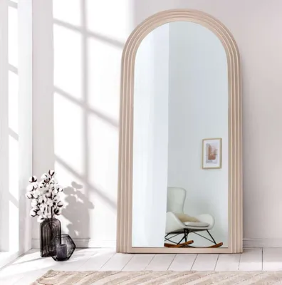 A full-length arched mirror with a fluted frame stands in a bright, minimalist room. Sunlight casts geometric shadows on the wall behind it. A vase of cotton branches is placed to the left of the mirror.