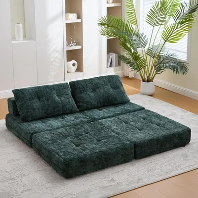 A modular green sofa is arranged on a light gray rug in a well-lit room with shelves and a large potted plant in the background.