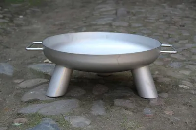 A metal fire pit with three legs and handles, sitting on a stone-covered ground.