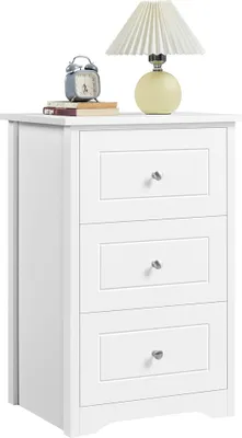 A white nightstand with three drawers is shown against a white background. On top of the nightstand are two books, a silver alarm clock, and a table lamp with a pleated shade and a yellow base.