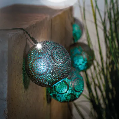 A string of solar-powered decorative lights with spherical, perforated shades in a teal and silver color scheme, hanging near a stone wall and tall grass.