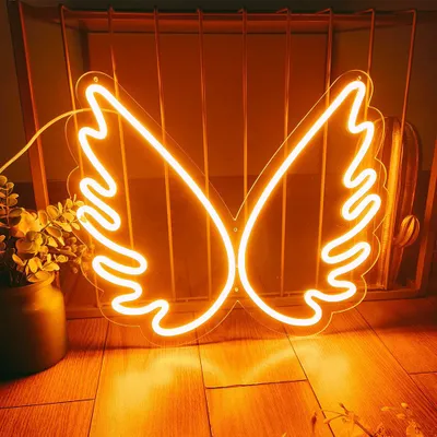 An orange neon angel wing sign is displayed on a wooden floor in front of a metal rack. A potted plant with yellow flowers is to the left of the sign.