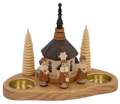 Ein hölzerner Teelichthalter zeigt eine Kirche umgeben von drei Figuren und zwei Bäumen auf einem ovalen Sockel mit zwei Teelichthaltern.