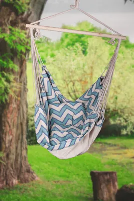 A hammock chair with a chevron pattern is hanging from a wooden frame in a garden setting with trees and grass.