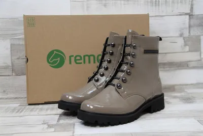 A pair of beige patent leather lace-up boots are displayed in front of their cardboard packaging.