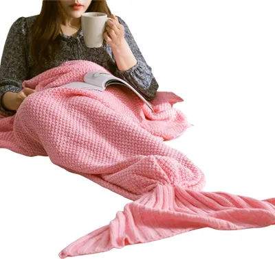 A person is sitting wrapped in a pink mermaid tail blanket, holding a mug and reading a book.