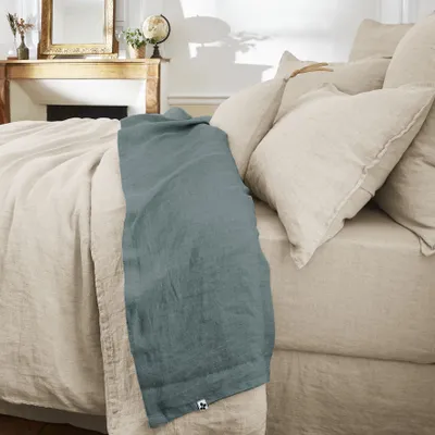 A bed with a beige duvet and pillows, with a blue throw blanket draped over it. A fireplace and mirror are visible in the background.