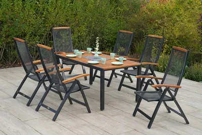 A patio dining set with a wooden table and six chairs, set up on a light-colored deck, with lush green foliage in the background.
