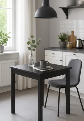 A small black dining table with a gray chair and a mug on it, set in a bright kitchen with a window and cabinets.