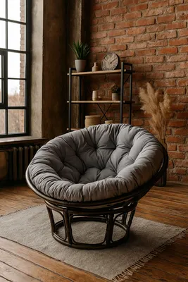 A papasan chair with a gray cushion sits on a rug in a room with exposed brick walls and a large window.