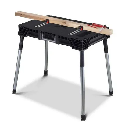A black folding workbench with silver legs is shown with a piece of wood clamped to the top.