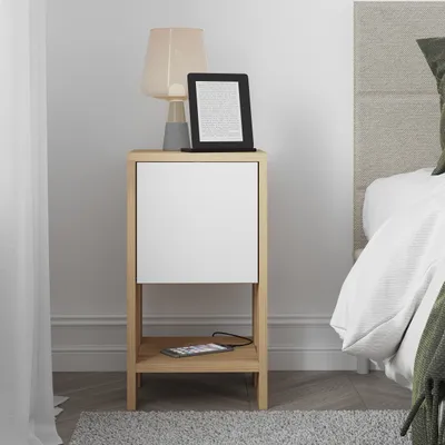 A bedside table with a drawer and an open shelf, placed next to a bed. A lamp and an e-reader are on top of the table, and a smartphone is on the shelf.