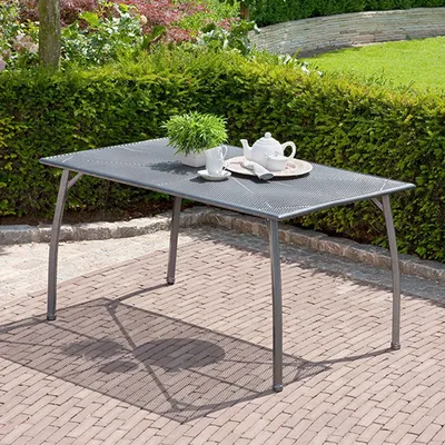 A metal garden table set with a teapot and cup on it, placed on a brick patio surrounded by greenery.