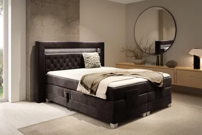 A bedroom scene featuring a dark brown upholstered box spring bed with an LED headboard, a white mattress, and beige bedding. A round mirror and a wooden dresser are visible in the background.