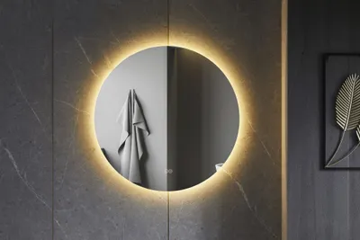 A round, frameless mirror with LED lighting is mounted on a dark gray tiled wall. A gray towel is hanging on the wall reflected in the mirror.