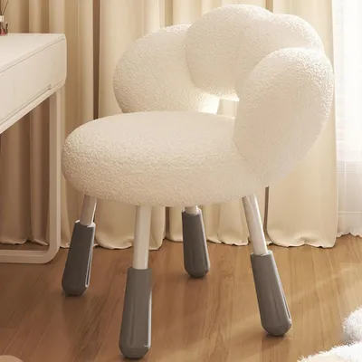 A cream-white fluffy chair with gray leg caps sits on a wooden floor next to a white desk and beige curtains.
