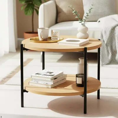 A round, two-tiered coffee table with a wooden top and black metal legs, placed in a living room setting with a sofa and plant in the background.