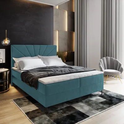 A bedroom scene featuring a teal boxspring bed with white bedding, a dark gray blanket, and a patterned rug.