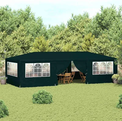 A dark green party tent set up in a grassy backyard with trees in the background. The tent has arched windows and an open entrance revealing tables and chairs inside.