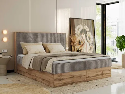 A bedroom scene featuring a large boxspring bed with a tall headboard, dressed with bedding and pillows. A large framed artwork leans against the wall behind the bed, and a plant is visible near a window.