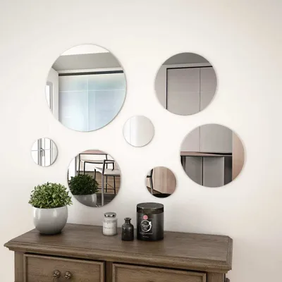 A collection of round mirrors of various sizes are displayed on a white wall above a wooden dresser. A potted plant and some decorative jars are on the dresser.