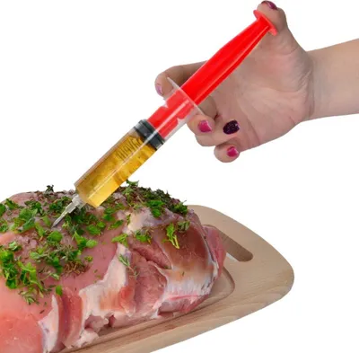 A hand is using a red and clear syringe to inject liquid into a piece of raw meat seasoned with herbs.