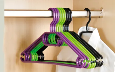 A group of plastic hangers in purple, green, and black are hanging on a metal closet rod. A white shirt is also visible on a black hanger in the background.