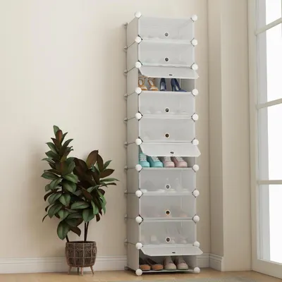A tall, narrow shoe rack with multiple compartments, some of which are open and contain shoes. A potted plant is to the left of the shoe rack.