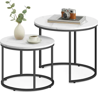Two nesting round coffee tables with marble-look tops and black metal legs, one with a vase of greenery and a cup of coffee, the other with a stack of books and a vintage camera.