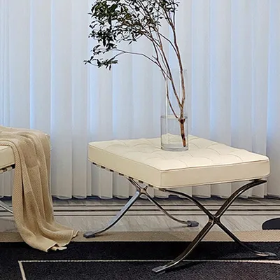 A beige leather ottoman with a tufted top and chrome legs sits in a room with sheer white curtains in the background.