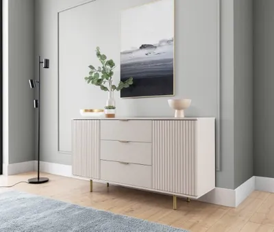 A modern sideboard with fluted doors and drawers, set against a light gray wall with a framed abstract painting above it. A floor lamp stands to the left, and a blue rug is visible in the foreground.
