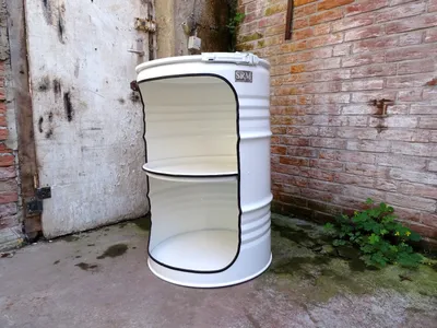 A white barrel-shaped shelf unit with two shelves sits on a concrete floor next to a brick wall and a weathered door.