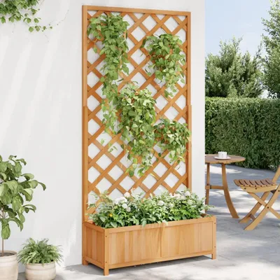 A wooden planter box with a trellis attached, filled with plants, stands against a white wall on a patio. A small round table and chair are visible in the background.