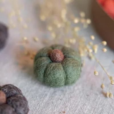 A small, green felt pumpkin with a brown stem sits on a textured surface, with blurred dried plant stems in the background.