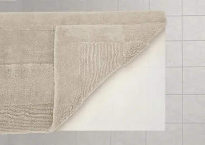 A beige bath mat is shown with one corner lifted, revealing a white backing. The mat is placed on a tiled floor.