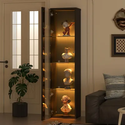 A tall, narrow display cabinet with glass doors and shelves illuminated by warm LED lights, showcasing several decorative busts and figurines. The cabinet is positioned in a room next to a doorway and a large potted plant, with a sofa visible in the backgr
