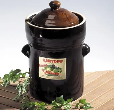 A brown ceramic fermentation crock with a lid, placed on a wooden surface with green ivy vines.