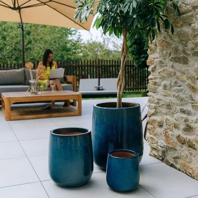 A woman is working on a laptop on a patio next to a sofa, with large blue planters and a tree in the foreground.