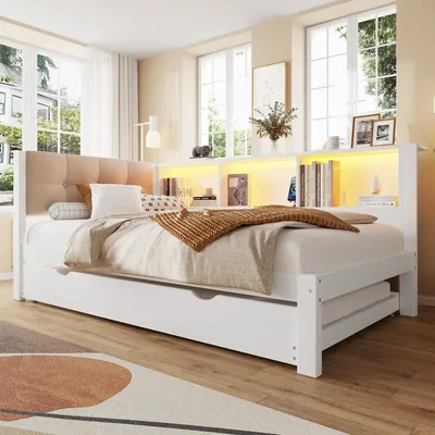 A bedroom scene featuring a white daybed with a tufted beige headboard, a pull-out trundle bed, and a built-in shelf unit with LED lighting.