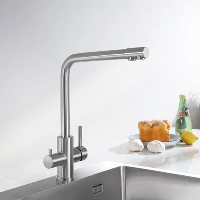 A stainless steel kitchen faucet is shown in a studio setting, with a sink and countertop visible. A bottle of water, a lemon, and two garlic bulbs are placed on the countertop.