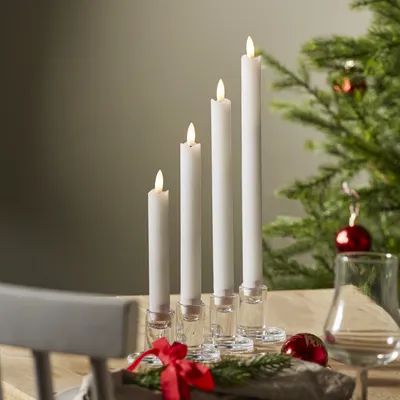 Four LED candles of varying heights are arranged on a table, with a Christmas tree and ornaments in the background.