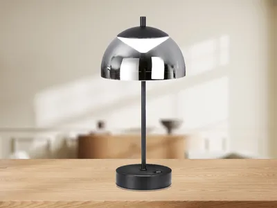 A modern black table lamp with a reflective silver dome shade is placed on a wooden table in front of a blurred interior background.