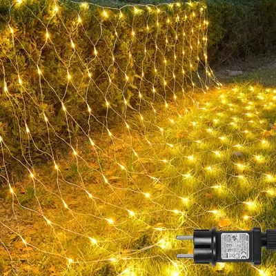 A net of warm white lights is draped over bushes and spread across the grass in a garden at dusk.