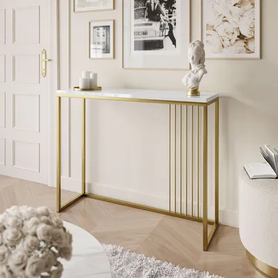 A console table with a white top and gold frame is placed in a well-decorated room with artwork on the wall and a coffee table in the foreground.