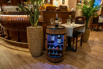 A wine barrel shaped cabinet with illuminated shelves, displaying wine bottles and glasses on top, in a restaurant setting.