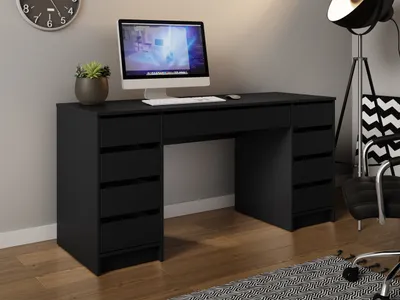 A black desk with a computer and a plant on it, in a room with a clock on the wall and a floor lamp and chair in the background.