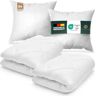 A set of white pillows and a duvet are arranged on a white background. The pillows are stacked in the foreground, and two larger pillows are in the background. One of the background pillows has a green band with text and logos.