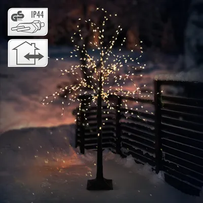 A decorative LED tree with warm white lights stands in a snowy outdoor setting at dusk, with a wooden fence in the background.