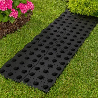 A black pathway mat is laid on a grassy lawn next to a bush and pink flowers.