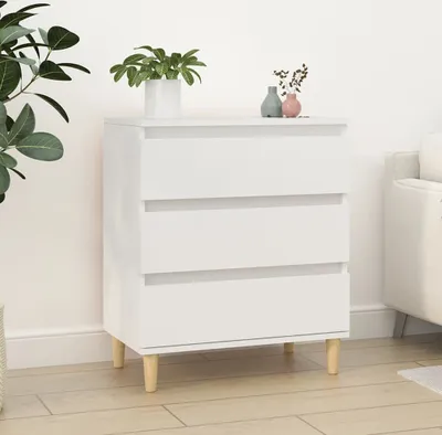 A white three-drawer dresser with wooden legs, placed against a white wall with a plant and two small vases on top.